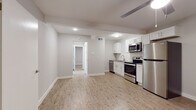 This single-floor unit showcases a thoughtfully updated interior with a cohesive, modern design throughout. Light wood-style luxury vinyl plank flooring runs continuously across all rooms, creating a warm and unified feel. The open kitchen is well-appointed with white shaker-style cabinetry, grey countertops, and a full suite of stainless steel appliances including a range, over-the-range microwave, dishwasher, and refrigerator. A white subway tile backsplash adds a clean, classic detail to the cooking area, which flows directly into an open living space with a ceiling fan overhead.

A hallway leads to the additional rooms, each finished with the same light flooring and neutral grey walls. One bedroom features a ceiling fan with a light fixture and a window that brings in natural light. A second room offers a versatile, open layout suitable for various uses. The bathroom is finished with large-format marble-style tile lining the tub surround, complemented by wood-look flooring, a white vanity, and brushed metal hardware. Throughout the unit, recessed lighting and clean white trim contribute to a bright, well-maintained atmosphere.