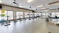 This single-floor fitness center is thoughtfully designed to accommodate a wide range of workout styles and training needs. The space is divided into distinct functional zones, each outfitted with appropriate equipment and finishing details to support an effective exercise environment.

The cardio area features multiple treadmills and stationary cycling bikes arranged along a wall lined with large windows fitted with horizontal blinds. Natural light filters in generously, offering views of the surrounding exterior. Ceiling fans and flush-mount fluorescent lighting provide consistent illumination and airflow throughout. A full-length mirror runs along one wall, visually expanding the space and allowing users to monitor their form.

The strength training zone includes a weight bench, a rack of dumbbells in graduated sizes, and additional step platforms. An accent wall in warm orange adds a bold visual contrast to the otherwise neutral beige palette. Ceiling fans continue throughout this area, maintaining comfort during intensive workouts.

A dedicated group exercise studio offers open floor space ideal for stretching, aerobics, or class-based training. Stability balls, spin bikes, and medicine balls are stored along the walls, and multiple windows ensure the room remains bright and well-ventilated. The durable, smooth flooring throughout the entire facility is well-suited to high-traffic fitness use.