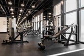Located at 200 City Blvd W in Orange, California, this fitness center offers a comprehensive workout environment designed to accommodate a wide range of training needs. The facility spans a generous single floor with an open, industrial aesthetic defined by an exposed structural ceiling with dark-painted beams, ductwork, and grid-pattern overhead framing, all illuminated by recessed and pendant lighting that creates a focused, energetic atmosphere.

The strength training area is well-appointed with a full rack of dumbbells, multiple weight benches, and a variety of cable and resistance machines, all arranged to maximize floor space and traffic flow. Full-length mirrors line the walls throughout, enhancing the sense of openness while allowing users to monitor their form. A dedicated cardio zone features stationary spin bikes, treadmills, and elliptical machines positioned near large windows that bring in natural light and views of the surrounding urban environment.

A separate functional training area provides open floor space for dynamic movement, complemented by medicine balls, a captain's chair, and additional cable machines. Durable rubber flooring throughout the facility ensures safety and comfort. A front service counter area and private rooms add further utility. The overall ambiance is modern, clean, and professionally outfitted, making it a fully functional fitness destination.