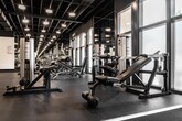 Located at 200 City Blvd W in Orange, California, this fitness center offers a comprehensive workout environment designed to accommodate a wide range of training needs. The facility spans a generous single floor with an open, industrial aesthetic defined by an exposed structural ceiling with dark-painted beams, ductwork, and grid-pattern overhead framing, all illuminated by recessed and pendant lighting that creates a focused, energetic atmosphere.

The strength training area is well-appointed with a full rack of dumbbells, multiple weight benches, and a variety of cable and resistance machines, all arranged to maximize floor space and traffic flow. Full-length mirrors line the walls throughout, enhancing the sense of openness while allowing users to monitor their form. A dedicated cardio zone features stationary spin bikes, treadmills, and elliptical machines positioned near large windows that bring in natural light and views of the surrounding urban environment.

A separate functional training area provides open floor space for dynamic movement, complemented by medicine balls, a captain's chair, and additional cable machines. Durable rubber flooring throughout the facility ensures safety and comfort. A front service counter area and private rooms add further utility. The overall ambiance is modern, clean, and professionally outfitted, making it a fully functional fitness destination.