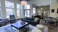 This single-floor space makes a strong first impression with its dramatic ceiling height and expansive windows that flood the interior with natural light. The main lounge area is anchored by a sleek charcoal sofa accented with green throw pillows, paired with a tufted leather ottoman coffee table and coordinating accent seating, including a patterned wingback chair and tufted ottomans. A statement drum pendant light hangs overhead, adding a touch of refined elegance to the gathering space. Dark hardwood flooring runs throughout, grounding the neutral gray wall palette and creating a warm, contemporary ambiance. Large floor-to-ceiling windows and a set of glass French doors open the space visually to lush outdoor greenery, enhancing the sense of openness and connection to the exterior. Adjacent to the lounge, a well-appointed kitchenette features dark wood cabinetry, light countertops, and modern appliances, making the space fully functional for hosting or everyday use. A small dining area completes the layout, offering a versatile setting suitable for both casual gatherings and productive meetings. Overall, the space balances modern design with practical amenities in a bright, welcoming environment.