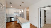 Located in Birmingham, AL, this single-floor unit offers a thoughtfully designed layout with modern finishes throughout. The kitchen serves as a centerpiece of the space, featuring a large central island topped with speckled granite countertops, pendant lighting with gold-toned hardware, and a subway tile backsplash. White upper cabinetry is paired with gray lower cabinets, and a full suite of stainless steel appliances — including a refrigerator, range, microwave, and dishwasher — adds a sleek, contemporary feel. Warm hardwood-style flooring extends through the kitchen and living areas, creating a cohesive and inviting atmosphere. The bedrooms are carpeted for comfort and are bright and airy, with at least one room featuring large windows fitted with white blinds that allow ample natural light. The bathroom is well-appointed with a granite-topped vanity, an undermount sink, a large frameless mirror, and hardwood-style flooring that continues the home's consistent design language. A louvered interior door adds a distinctive architectural touch visible throughout the unit. Clean white walls, recessed lighting, and modern fixtures contribute to the overall contemporary ambiance of this well-finished Birmingham residence.