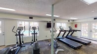 This fitness center in Lafayette, Louisiana, offers a practical and well-organized space designed to accommodate a variety of workout routines. The single-level layout is finished with patterned carpet tiles throughout the main exercise area, neutral-toned walls, and a textured ceiling with recessed fluorescent panel lighting that ensures consistent illumination across the space.

The main workout area is generously sized and houses a selection of cardio equipment, including multiple Star Trac treadmills, elliptical trainers, and stationary bikes, both upright and recumbent. A multi-station cable weight machine is positioned along one wall, providing options for strength training. Wall-mounted flat-screen televisions are positioned for easy viewing during cardio sessions. Multiple windows along the perimeter allow natural light to fill the room and offer views of an outdoor pool area, contributing to an open and energizing atmosphere. A glass-paneled door provides direct access to the exterior.

The facility also includes a restroom featuring tile flooring, a dark wood vanity with a white countertop sink, a framed mirror, and tile wainscoting. Near the entry area, a sanitation station with paper towel dispensers and a waste bin supports general cleanliness throughout the space.