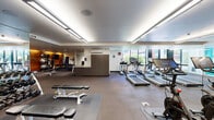 Located at 3750 S River Pkwy in Portland, Oregon, this well-appointed fitness center offers a comprehensive and modern workout environment spread across a single, generously sized floor. The space is thoughtfully designed with distinct functional zones to accommodate a wide range of exercise activities.

The cardio area is anchored by a row of True-brand treadmills, elliptical trainers, upright and recumbent stationary bikes, and stair climbers, all positioned to take advantage of large windows and glass doors that flood the space with natural light and provide views of the surrounding outdoor landscape. A set of French doors opens directly to an outdoor terrace area, adding a bright, airy ambiance to the room.

The free weights section features a full dumbbell rack, adjustable benches, and various strength training accessories, complemented by floor-to-ceiling mirrors that enhance the sense of openness. A warm wood accent wall adds a distinctive architectural touch to this zone.

A separate glass-walled studio area with a vibrant teal accent panel provides a dedicated space for stretching, cycling, or group workouts. The flooring throughout consists of durable rubber tiles, and the space is climate-controlled via wall-mounted HVAC units. Recessed and linear LED lighting ensures consistent, well-distributed illumination. A bathroom is also included within the facility.