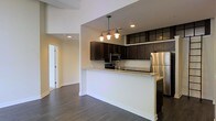 This single-floor space is designed with a clean, contemporary aesthetic that emphasizes an open and functional layout. The kitchen serves as a central highlight, featuring rich dark wood cabinetry that extends to the upper walls, providing ample storage. Granite countertops add an elegant touch, while the peninsula breakfast bar creates a natural divide between the kitchen and the adjacent living area, making it ideal for casual dining or entertaining. Stainless steel appliances, including a refrigerator, microwave, and range, lend a polished, modern feel to the space. A stylish multi-bulb pendant light fixture hangs above the bar, adding both warm illumination and a decorative focal point. Recessed lighting throughout the ceiling further brightens the interior. The flooring is rich, dark hardwood, which complements the cabinetry and flows seamlessly through the open-plan living area. Neutral, light-toned walls keep the space feeling bright and airy, offering a versatile backdrop for a variety of design styles. A hallway visible in the background leads to additional rooms, suggesting a well-organized and practical floor plan. Overall, the space balances style and utility in a cohesive, inviting manner.