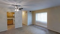 This single-floor residence offers a straightforward and functional layout designed for comfortable everyday living. The main living area is carpeted throughout, providing a warm and soft underfoot feel, and is illuminated by a ceiling fan fitted with integrated lighting. A large window dressed with horizontal blinds allows natural light to fill the room, creating a bright and airy atmosphere. The neutral, light-toned walls contribute to the open feel of the space, making it easy to personalize with a variety of décor styles. A closet with a classic paneled white door provides convenient built-in storage directly off the living area. HVAC vents are positioned in the ceiling and wall, ensuring climate control throughout the space. Adjacent to the living area, a glimpse of the kitchen reveals cabinetry and appliances, including what appears to be a range and refrigerator, suggesting a compact yet practical cooking space. The transition between the living and kitchen areas is seamless, supporting an open and connected flow. Overall, the residence presents a clean, modest, and well-maintained interior suited for functional day-to-day living.