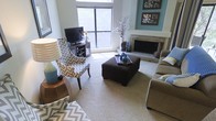 This single-story living space showcases a well-appointed living room that balances comfort and style. A standout fireplace with a light stone surround serves as a focal point, flanked by a teal accent wall adorned with decorative artwork. Large windows allow abundant natural light to fill the room, creating a bright and airy atmosphere with views of surrounding greenery. The space features plush carpeting underfoot, contributing to a warm and inviting ambiance. Seating is arranged thoughtfully around a dark brown ottoman, which serves as a central coffee table, complemented by a chevron-patterned accent chair and a neutral-toned sofa with decorative throw pillows. A media console positioned near the windows accommodates a flat-screen television, while a decorative vase with branches adds a natural, organic touch to the room. The teal and gray color scheme is carried throughout the space through carefully selected accessories and furnishings, creating a cohesive and polished aesthetic. Overall, the living room presents a functional yet stylish environment suited for both relaxation and entertaining.