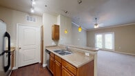 Located at 6805 Lebanon Rd in Frisco, Texas, this single-floor residence offers a thoughtfully designed layout that balances comfort and functionality. The kitchen is a standout feature, equipped with warm-toned wooden cabinetry, light countertops, stainless steel appliances including a full-size refrigerator, built-in microwave, and dishwasher. Rich wood-look flooring grounds the kitchen space, while amber pendant lights add a warm, inviting glow over the peninsula breakfast bar. The kitchen opens seamlessly into the living area, which is finished with plush carpeting, crown molding, and elevated ceilings that enhance the sense of spaciousness. A ceiling fan provides comfort, and French doors lead to a private balcony, filling the room with natural light. Adjacent to the living area, a built-in shelving nook offers practical storage or display space. The bedroom features wall-to-wall carpeting and a generously sized window fitted with blinds, allowing for flexible natural light control. The overall color palette of warm beige tones throughout creates a cohesive, neutral ambiance. The layout, as seen in the dollhouse view, is efficiently organized to maximize flow between the kitchen, living, and sleeping areas.