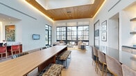 This thoughtfully designed single-floor communal space spans a generous footprint, seamlessly blending modern and warm design elements throughout its distinct functional zones. The main lounge and gathering area commands attention with soaring ceilings clad in rich wood paneling, complemented by ambient LED cove lighting that adds a warm, inviting glow. A dramatic wall of floor-to-ceiling steel-framed windows floods the space with abundant natural light and offers views of the surrounding exterior. A long communal table with patterned bench seating serves as a central workspace, while a dedicated workstation area with red accent chairs and a textured wall installation provides additional productivity options. Terracotta brick flooring flows throughout much of the space, adding texture and warmth, while large-format tile defines the main gathering areas. A columned corridor lined with curated artwork leads to an intimate lounge corner featuring a brick fireplace, built-in display shelving, a wall-mounted television, and a coffered wood-beam ceiling. Deep navy blue accent walls in this area create a cozy, distinct atmosphere. Leather and velvet seating arrangements throughout offer multiple opportunities for comfortable social interaction, making this a versatile and visually rich communal environment.