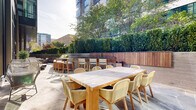 Located at 44 M St NE in Washington, DC, this elegantly designed outdoor terrace offers a refined urban retreat in the heart of the city. The space is finished with large-format stone pavers in a rich, veined grey palette, providing a polished and durable foundation throughout. Floor-to-ceiling glass walls along one side of the terrace connect the outdoor environment seamlessly to the interior, flooding both spaces with abundant natural light.

The terrace is thoughtfully divided into distinct functional zones. A generous communal dining area features a solid wood teak table paired with woven rattan chairs, ideal for group gatherings or al fresco meals. Adjacent to this, a bar-height counter with teak stools provides a more casual seating option bathed in sunlight. Cushioned lounge chairs and rounded accent tables create intimate conversation areas perfect for relaxed socializing.

Anchoring the perimeter is a continuous built-in cedar bench with integrated raised planters overflowing with lush greenery and seasonal plantings. Dense hedgerows and mature trees further enclose the space, offering privacy and a strong sense of seclusion within the urban setting. The surrounding mid-rise Washington, DC skyline is visible above the treeline, reinforcing the terrace's vibrant city context while maintaining a calm, garden-like atmosphere.