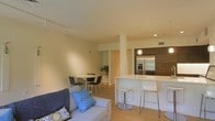 This stylish single-floor space showcases a thoughtfully designed open-concept layout that effortlessly connects the living, dining, and kitchen areas into one cohesive, functional environment. The living area is anchored by a comfortable sofa accented with decorative throw pillows, complemented by warm, light-toned flooring that runs throughout the space and contributes to a bright, airy atmosphere. Recessed lighting and a track lighting system illuminate the room evenly, enhancing the clean, modern aesthetic.

The kitchen is a standout feature, boasting sleek, dark wood cabinetry, a stainless steel French door refrigerator with a built-in freezer drawer, and a wall oven. A crisp white kitchen island with bar-height seating provides both additional prep space and a casual dining option, making it ideal for entertaining. A pendant light adds a refined touch above the island area.

Adjacent to the kitchen, the dining area accommodates a round table with modern chairs, creating an intimate yet open setting for meals. Structural columns add architectural character while subtly defining the different zones within the open floor plan. The overall ambiance is contemporary and welcoming, with neutral tones and clean lines throughout that create a calm, livable environment.