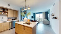 Located along the desirable North Bayshore Drive corridor in Miami, FL, this thoughtfully designed single-floor residence blends contemporary style with coastal-inspired living. The open-concept kitchen and living area serves as the heart of the home, anchored by a warm wood-toned island with a white quartz countertop and a striking brass pendant light fixture overhead. Custom cabinetry, stainless steel appliances, and a tiled backsplash complete the well-appointed kitchen. Bold teal accent walls and large sliding glass doors in the living area flood the space with natural light and provide direct access to a private balcony with sweeping outdoor views of the Miami skyline. Multiple bedrooms offer distinct design personalities — one features deep navy walls and soft sheer drapes framing outdoor views, another presents a bright, neutral palette with floor-to-ceiling windows. Each bedroom benefits from abundant natural light and scenic vistas. The bathroom is finished with large-format floor tiles, a dual-sink wood vanity with quartz countertops, a glass-enclosed shower, and a rich burgundy accent wall. Large tile flooring flows consistently throughout, tying together the home's clean, modern aesthetic.