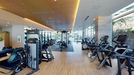 This well-appointed fitness center is thoughtfully designed to accommodate a comprehensive range of workout disciplines within a single, expansive floor. The space is divided into distinct functional zones, including a dedicated cardio area, a strength training section, and a functional training zone, each seamlessly connected by light hardwood flooring and rubber-matted surfaces underfoot.

Floor-to-ceiling glass walls line the perimeter, flooding the interior with abundant natural light while offering views of lush palm trees and landscaped outdoor surroundings. This transparency creates an open, airy atmosphere that enhances the overall workout experience. The warm wood-paneled ceiling, accented by recessed amber uplighting, adds a refined, boutique-hotel quality to the space.

The cardio zone houses a full lineup of True treadmills, ellipticals, stair climbers, and stationary bikes, all positioned to maximize the outdoor views. The strength area features Cybex Eagle selectorized machines and a comprehensive free weight station with a full dumbbell rack. A functional training area with TRX suspension systems, balance equipment, and plyometric boxes rounds out the facility. Stone-clad structural columns and full-length mirrors contribute to the polished, modern aesthetic throughout.