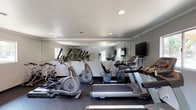 This single-floor fitness center is thoughtfully designed to provide a functional and motivating workout environment. The space is outfitted with a variety of cardio equipment, including treadmills, elliptical trainers, and stationary cycling bikes, offering users multiple options for cardiovascular exercise. A dedicated free weight area features a full rack of dumbbells in graduated sizes, catering to strength training needs. Large mirrored walls span a significant portion of the room, visually expanding the space and allowing users to monitor their form during workouts. A wall-mounted flat-screen television adds an entertainment element to the cardio experience. The flooring consists of durable rubber matting, providing both safety and comfort underfoot. Recessed lighting in the white ceiling ensures the space is well-lit throughout, while natural light filters in through windows along the walls, creating a bright and energizing atmosphere. HVAC vents integrated into the ceiling maintain a comfortable climate during workouts. The overall design is clean, practical, and efficiently organized to maximize usable floor space for a variety of fitness activities.