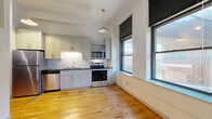 Located at 1 E First St in the heart of downtown Duluth, Minnesota, this efficiently designed single-floor unit blends urban character with modern updates. Original hardwood floors run throughout the main living areas, lending warmth and a nod to the building's historic roots. Soaring ceilings amplify the sense of openness, while a ceiling fan and pendant lighting add both function and style.

The kitchen is a standout feature, thoughtfully outfitted with light gray shaker-style cabinetry, dark countertops, and a classic white subway tile backsplash. A full suite of stainless steel appliances — including a refrigerator, range, over-the-range microwave, and dishwasher — makes it as practical as it is attractive. Large windows along the kitchen wall invite natural light and offer views of the surrounding cityscape.

Dark-stained barn-style sliding doors define the entry corridor, adding an industrial-chic aesthetic that complements the building's character. An in-unit washer is also present, adding everyday convenience.

The bathroom is clean and contemporary, featuring a white vanity with brushed nickel hardware, a large mirror, tile flooring, and a full shower-tub combo. The overall ambiance of the space is bright, modern, and functional — an ideal urban retreat in the heart of Duluth.
