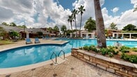 Located at 900 Downtowner Boulevard in Mobile, Alabama, this outdoor amenity area presents a resort-inspired pool environment designed for relaxation and recreation. The centerpiece is a generously sized, freeform swimming pool with crystal-clear blue water, accessible via stainless steel entry rails and surrounded by an expansive decorative paver deck featuring a distinctive geometric star pattern throughout.

Ample seating is provided by numerous blue lounge chairs positioned along the pool's perimeter, offering sun-drenched spots for leisure. Towering palm trees are strategically placed around the area, providing natural shade and lending a distinctly tropical character to the space. Raised stone planter beds add visual interest and texture, filled with lush cycad palms and tropical foliage that enhance the resort ambiance.

A white columned pergola serves as an elegant architectural focal point, creating a shaded gathering area alongside the pool deck. A charming brick cabana structure with a hip roof is also present, adding functional utility to the outdoor space. Well-maintained landscaping with manicured shrubs and mature trees borders the entire area, providing both privacy and a verdant backdrop. The open-sky setting allows for abundant natural light, completing the inviting, amenity-rich outdoor experience.