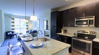 This single-floor residence showcases an open-concept design that integrates the kitchen, living, and sleeping areas into one cohesive, flowing space. The kitchen serves as the focal point, anchored by a large island with striking light-speckled granite countertops that provide generous prep and dining space. A stainless steel undermount sink is centrally positioned on the island, accompanied by a sleek pull-down faucet. Pendant lights with brushed metal fixtures hang above, casting warm, focused illumination across the workspace. Dark espresso-toned cabinetry lines the perimeter walls, extending floor to ceiling to maximize storage capacity, and is complemented by stainless steel appliances, including a built-in microwave and range. The transition from the kitchen to the living area is seamless, where natural light filters in through large windows, offering a bright and airy ambiance. A glimpse into the bedroom beyond reveals continued natural light and a cohesive design aesthetic throughout. The overall space balances contemporary finishes with functional layout, creating an environment that feels both modern and welcoming.