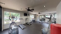 This single-level fitness center offers a comprehensive workout environment designed to support a wide range of exercise routines. The space is finished with durable rubber flooring throughout, providing a safe and practical surface suited for both cardio and strength training activities. Recessed lighting and multiple ceiling fans are installed across the ceiling, ensuring consistent illumination and airflow during workouts.

Cardio equipment includes treadmills and an elliptical trainer, positioned to take advantage of the natural light streaming in through several large windows. These windows also offer views of a surrounding pool area and landscaped grounds, creating an inviting atmosphere. A wall-mounted television is positioned for easy viewing during exercise.

The strength training section is well-appointed, featuring a multi-station cable machine, adjustable bench press, vertical knee raise station, and a tiered dumbbell rack stocked with a varied selection of free weights. Kettlebells, stability balls, battle ropes, and medicine balls are also available, supporting functional and cross-training workouts.

Full-length mirrors line one wall, visually expanding the space and assisting with form correction. Large glass doors connect the fitness center to an adjacent common area, enhancing the sense of openness and allowing abundant natural light to flow through the interior.