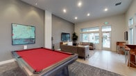 This contemporary amenity lounge is designed to offer a well-rounded recreational and social environment across a single, open floor. The space is anchored by a striking red felt pool table positioned at the center, set atop a large area rug that defines the entertainment zone. Soaring ceilings and an abundance of large windows and glass doors flood the interior with natural light while providing direct views of and access to an outdoor pool area.

The lounge area features a tufted leather sofa, a live-edge coffee table, and a wall-mounted flat-screen TV above a mid-century modern media console, creating a comfortable and stylish gathering spot. A tall indoor plant adds a touch of greenery to the room.

Adjacent to the main lounge is a compact dining nook with high-top tables and modern bar-height chairs positioned beneath large street-facing windows, ideal for casual seating or work. The fully equipped kitchenette showcases warm wood cabinetry, a bold geometric backsplash tile, a stainless steel refrigerator, a sink, and a white quartz island with bar seating — well-suited for hosting events or everyday use.

Throughout the space, large-format tile flooring, recessed lighting, and a neutral gray and white color palette contribute to a clean, modern aesthetic that is both functional and inviting.
