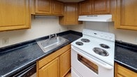 This single-floor residential unit offers a straightforward and functional layout designed for comfortable everyday living. The kitchen is equipped with warm oak cabinetry, dark granite-style countertops, a stainless steel sink, and a white electric range with overhead ventilation, providing a practical space for meal preparation. The living and bedroom areas are finished with wall-to-wall carpeting in neutral tones, creating a soft and cohesive feel throughout. The bedrooms benefit from natural light streaming through blinded windows that frame views of surrounding greenery, lending a calm and inviting atmosphere to each room. The spacious main living area features a solid wood entry door, textured ceilings, and wainscoting detail that adds a subtle architectural touch. The unit includes two bathrooms: one featuring a full soaking tub with white subway-tile surround and chrome fixtures, and a second with a wide white countertop vanity, an integrated sink, and a large mirror with built-in storage and overhead lighting. Tile flooring in the bathrooms contrasts neatly with the carpeted living spaces. The overall palette of warm whites and beiges throughout creates a clean, neutral canvas well-suited to a variety of interior styles.