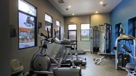 This fitness center offers a thoughtfully designed workout space that balances functionality with a modern aesthetic. The interior is finished with neutral gray walls and recessed lighting that work in harmony to create a bright, energizing atmosphere. Large windows line the walls, allowing ample natural light to fill the space while offering views of the surrounding outdoor area, including a glimpse of a pool and landscaped grounds beyond.

The facility is outfitted with a comprehensive selection of Matrix-brand cardio equipment, including elliptical trainers and treadmills, arranged to maximize floor space and ease of movement. A multi-station cable and strength training machine is positioned along one side, providing users with a versatile range of resistance exercise options. The durable, textured flooring throughout is well-suited for high-traffic gym use and easy maintenance.

Recessed ceiling vents ensure proper ventilation, while safety features such as clearly marked exit signage contribute to a well-organized environment. The single-floor layout makes the entire facility easily accessible, offering a streamlined experience for users of all fitness levels. Overall, this fitness center presents a clean, professional, and fully functional space for cardiovascular and strength training workouts.
