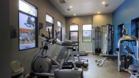 This fitness center offers a thoughtfully designed workout space that balances functionality with a modern aesthetic. The interior is finished with neutral gray walls and recessed lighting that work in harmony to create a bright, energizing atmosphere. Large windows line the walls, allowing ample natural light to fill the space while offering views of the surrounding outdoor area, including a glimpse of a pool and landscaped grounds beyond.

The facility is outfitted with a comprehensive selection of Matrix-brand cardio equipment, including elliptical trainers and treadmills, arranged to maximize floor space and ease of movement. A multi-station cable and strength training machine is positioned along one side, providing users with a versatile range of resistance exercise options. The durable, textured flooring throughout is well-suited for high-traffic gym use and easy maintenance.

Recessed ceiling vents ensure proper ventilation, while safety features such as clearly marked exit signage contribute to a well-organized environment. The single-floor layout makes the entire facility easily accessible, offering a streamlined experience for users of all fitness levels. Overall, this fitness center presents a clean, professional, and fully functional space for cardiovascular and strength training workouts.