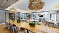 Located at 4180 Sunrise Hwy in Oakdale, NY, this thoughtfully designed single-floor space seamlessly blends a dining area and living room into one cohesive, open-concept environment. The interior showcases a refined coastal aesthetic, anchored by warm herringbone-patterned hardwood flooring, cove lighting, and a distinctive slatted wood ceiling detail that adds texture and depth.

The dining area is a standout, featuring a striking live-edge communal table paired with woven rattan pendant lights that serve as sculptural focal points overhead. Multiple dining tables with upholstered chairs in neutral tones offer flexible seating arrangements, while a bar-height counter with stools provides an additional casual dining option. Floral drapery panels frame large French doors that open to an outdoor patio, flooding the space with natural light and views of the surrounding exterior.

The living area is anchored by a generously sized sectional sofa arranged around a wall-mounted flat-screen television set against a stone-textured accent wall. Bold pendant lights with a matte black and gold interior add a modern contrast to the otherwise soft, neutral palette. Lush tropical potted plants are positioned throughout, enhancing the bright, airy ambiance. A transitional lounge area with a velvet settee and mirrored wall panels provides an elegant passage between the two zones.