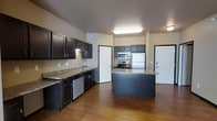 This single-floor space offers a thoughtfully designed interior that combines functionality with modern aesthetics. The kitchen is the centerpiece of the unit, showcasing rich dark espresso cabinetry that contrasts beautifully with granite-style countertops. A central island provides additional prep space and casual seating, making it ideal for both cooking and entertaining. Stainless steel appliances — including a refrigerator, range, microwave, and dishwasher — complete the kitchen's contemporary look. Warm hardwood flooring flows throughout the open layout, adding a sense of continuity and warmth to the space. The open-concept design allows for generous sightlines from the kitchen into the adjoining living areas, creating an airy and connected feel. Recessed and flush-mounted ceiling lighting ensures the space is well-lit throughout. Arched doorways add an architectural charm that softens the overall design. The neutral wall tones complement the dark cabinetry and flooring, resulting in a balanced and inviting ambiance that suits a variety of interior styles.