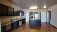 This single-floor space offers a thoughtfully designed interior that combines functionality with modern aesthetics. The kitchen is the centerpiece of the unit, showcasing rich dark espresso cabinetry that contrasts beautifully with granite-style countertops. A central island provides additional prep space and casual seating, making it ideal for both cooking and entertaining. Stainless steel appliances — including a refrigerator, range, microwave, and dishwasher — complete the kitchen's contemporary look. Warm hardwood flooring flows throughout the open layout, adding a sense of continuity and warmth to the space. The open-concept design allows for generous sightlines from the kitchen into the adjoining living areas, creating an airy and connected feel. Recessed and flush-mounted ceiling lighting ensures the space is well-lit throughout. Arched doorways add an architectural charm that softens the overall design. The neutral wall tones complement the dark cabinetry and flooring, resulting in a balanced and inviting ambiance that suits a variety of interior styles.