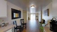 This single-floor space is designed with an open, flowing layout that connects the dining and living areas seamlessly. Hardwood-style flooring runs throughout, lending warmth and visual continuity to the interior. The dining area accommodates a set of black bar-style chairs paired with a compact table, complemented by a decorative framed mirror with a bold, textured border mounted on the wall nearby.

The living area extends toward the rear of the space, where a large sliding glass door with vertical blinds allows generous natural light to fill the room. A wall-mounted air conditioning unit is positioned near the sliding door, providing climate control. The neutral, light-toned walls and textured ceiling contribute to a clean, bright atmosphere throughout.

A hallway near the entrance leads to additional areas of the unit, with a door visible along its path. A small accent table with a green metal chair is positioned near the entrance, adding a functional yet decorative touch. Overall, the space presents a practical and well-lit interior with a straightforward, comfortable layout suited for everyday living.