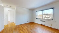 This single-floor unit efficiently accommodates four bedrooms, one bathroom, a living room, a kitchen, and a connecting hallway within a compact footprint. Warm-toned hardwood flooring runs consistently throughout the living areas and bedrooms, lending a cohesive and classic character to the interior.

Each bedroom is modestly sized, featuring white walls, hardwood floors, and individual windows that admit natural light and outdoor views of surrounding trees and neighborhood streetscapes. Radiator heating units are present beneath the windows in several rooms. The primary bedroom includes a ceiling fan and dual corner windows, enhancing both ventilation and brightness. Additional bedrooms offer closet space, including at least one with a hanging rod and open shelving.

The kitchen is equipped with a stainless steel gas range, wood cabinetry, tile flooring, and an electrical panel, providing functional utility in a compact layout. The bathroom features classic mosaic tile flooring in muted pink and blue tones, white wall tiles, a bathtub, toilet, and a window for natural ventilation. A hallway connects the rooms, facilitating straightforward navigation throughout the unit.