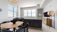 This single-floor space showcases a cohesive contemporary design with a neutral palette of whites and grays accented by warm wood tones. The open-plan living and dining area serves as the heart of the unit, where a generously sized sectional sofa in soft blue-gray velvet anchors the living space beneath a white-painted ceiling with exposed structural details. Large windows fitted with horizontal blinds draw in abundant natural light while offering views of the surrounding cityscape. A round wooden dining table with black chairs sits adjacent, creating a seamless transition between living and dining zones.

The kitchen is compact yet well-appointed, featuring flat-front cabinetry in a warm gray wood-grain finish, stainless steel appliances including a full-size refrigerator and range with hood, and light countertops with under-cabinet lighting.

Two bedrooms offer restful retreats, each furnished with a wood-framed bed dressed in crisp white bedding, wall-mounted reading lights, and blinds providing light control. The primary bedroom benefits from a generous window, a wall-mounted television, and a bench at the foot of the bed.

The bathroom is finished with fine mosaic tile flooring, a floating wood-grain vanity with a white countertop, a large mirror, and a glass-enclosed walk-in shower with gray tile surround, completing the space's polished, modern aesthetic.
