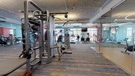 This spacious fitness center is organized into several distinct workout zones, each designed to support a comprehensive exercise experience. The main strength training area features a robust selection of cable machines, weight benches, and free weights from brands such as Hoist and Cybex, arranged along striped, multi-tone carpeted flooring. Bold accent walls in orange and red provide visual energy throughout the space, while large windows at the far end draw in natural light and offer outdoor views.

A separate glass-enclosed studio houses spin bikes and stability balls, illuminated by elegant circular pendant lighting and lined with mirrored walls to enhance the sense of space. This dedicated room features smooth hardwood-style flooring, suitable for cycling and group fitness classes.

The cardio zone lines the windowed perimeter, outfitted with stair climbers, ellipticals, and upright bikes facing mounted flat-screen televisions. A textured ceiling runs throughout the facility, paired with linear fluorescent and recessed lighting. A glass-walled entry corridor leads into the main floor, with a water fountain and waste station conveniently positioned near the entrance. Mirrors are strategically placed throughout to expand the visual footprint of the facility.