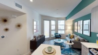 This thoughtfully designed single-floor residence combines contemporary style with functional living. The open-plan layout seamlessly connects the dining and living areas, creating a cohesive and inviting atmosphere. White walls provide a clean backdrop, punctuated by a striking teal accent wall that adds depth and personality to the living space.

The living area is anchored by large bay-style windows and a glass-paneled door that flood the interior with abundant natural light. These windows frame views of the surrounding urban environment while maintaining an airy, open feel. The flooring throughout is warm hardwood-style, complementing both the neutral tones and vibrant accents present in the space.

The dining area is positioned adjacent to the living room, with a clean white table providing a natural boundary between the two zones. A dark-toned credenza adds contrast and serves as practical storage near the entryway.

A private balcony accessed through the living area's glass door overlooks a well-appointed courtyard below, complete with a swimming pool, landscaping, and communal amenities — offering a pleasant urban retreat. The overall ambiance is modern, vibrant, and light-filled.
