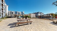 Located at 1880 Maple Dr NW in Kennesaw, Georgia, this outdoor communal space serves as a central gathering area within a modern multi-story residential community. The courtyard is finished with a broad, patterned stone paver surface that extends generously across the open-air area, providing a clean and durable foundation for a variety of recreational uses.

At the heart of the space sits a circular fire pit surrounded by cushioned outdoor seating, creating an inviting focal point for social gatherings. Multiple picnic tables with bench seating are distributed throughout the courtyard, offering ample space for dining and casual outdoor activities. Lush tropical plantings, including palm trees and ornamental shrubs, are strategically placed to add greenery and visual interest.

The courtyard is enclosed by a black iron perimeter fence with a gated access point, which includes a keypad entry system for resident security. Landscape beds line portions of the fence line, incorporating native plants and decorative ground cover. A community pool is visible just beyond the courtyard boundary, accessible through the gated entrance.

The surrounding multi-story buildings feature contemporary architecture with blue and gray façades, balconies, and large windows. The open-sky setting allows abundant natural light to fill the space throughout the day, making this outdoor area a well-equipped and attractive amenity for the Kennesaw community.