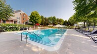 Located at 101 Cambridge Ln in Newtown, PA, this outdoor community pool facility offers a well-appointed recreational retreat designed for relaxation and leisure. The centerpiece is a generously sized swimming pool with crystal-clear blue water, accessible via built-in entry steps and a stainless steel handrail, making it both inviting and easy to enter. The pool is surrounded by a broad, open concrete sundeck that provides ample space for lounging and socializing.

The deck is furnished with numerous sling-style chaise lounges arranged along the pool's perimeter, offering residents plenty of options for sunbathing. Shaded seating areas with market umbrellas and accompanying tables provide comfortable alternatives for those seeking relief from direct sun. A wrought iron perimeter fence encloses the entire pool area, ensuring a safe and defined recreational zone.

Mature deciduous trees line the boundaries of the space, casting natural shade across portions of the deck and creating a lush, park-like atmosphere. Views beyond the fence reveal well-maintained green lawns and neighboring residential structures. A pool house or utility building is visible at one end of the enclosure, supporting the operational needs of the facility. The overall ambiance is calm, clean, and community-oriented, making this an ideal outdoor amenity for warm-weather enjoyment.