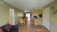 This single-floor residence showcases an open-concept design that seamlessly connects the living, dining, and kitchen areas into one cohesive space. Warm hardwood flooring runs throughout, complementing the neutral beige walls and light wood cabinetry to create a welcoming, unified aesthetic.

The kitchen is well-appointed with light wood cabinetry extending to the upper walls, a black electric range, a full-size refrigerator, and ample counter space along a peninsula that separates the kitchen from the dining area. The layout promotes functionality while maintaining visual openness.

Adjacent to the kitchen, the dining area accommodates a round table with wicker-style chairs, offering a casual yet comfortable setting. A ceiling fan overhead provides air circulation and lighting across the open space.

The living area flows naturally toward a glass sliding door or window at the far end, allowing natural light to filter through and brighten the interior. A textured popcorn ceiling adds a classic residential character, while framed artwork on the walls contributes a personal touch. The overall ambiance is casual, bright, and practical, suited for everyday living.