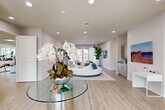 Located at 624 NE 2nd St in Dania Beach, Florida, this single-story amenity facility offers a thoughtfully designed and well-appointed interior spread across a spacious single level. Light wood-look flooring flows throughout, unifying the distinct functional zones and contributing to the space's clean, contemporary aesthetic.

The welcoming lounge area serves as the heart of the space, featuring a striking circular sectional sofa in white and blue, set atop a striped area rug. Bold, colorful abstract artwork adorns the walls alongside a wall-mounted flat-screen TV and a sleek navy blue credenza with gold accents. Tall tropical plants, sheer draping curtains, and glass-top accent tables complete the sophisticated yet inviting atmosphere. Floor-to-ceiling glass doors open to an outdoor patio, bathing the interior in abundant natural light and providing views of the surrounding landscape.

The fitness center is generously sized and fully equipped, featuring cardio machines including treadmills and a stair climber, strength-training stations, a full rack of dumbbells, workout benches, and a dedicated functional training area with stability balls and resistance equipment. Floor-to-ceiling mirrors amplify the sense of space, while multiple windows ensure bright, naturally lit workouts.

A clean, ADA-accessible corridor leads to restrooms and utility closets, rounding out this well-organized, modern facility.