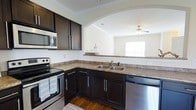 Located at 100 Durham Park Way in Pooler, Georgia, this two-story residence offers a thoughtfully designed layout that balances functionality with modern style. The kitchen is a standout feature, showcasing rich dark cabinetry with sleek hardware, granite-look countertops, and a full suite of stainless steel appliances, including an electric range, built-in microwave, and dishwasher. A double basin sink is positioned beneath a window, allowing natural light to fill the space. An elegant arched passageway connects the kitchen to the adjoining living area, creating an open and airy flow between the two spaces. Crown molding adds a refined architectural detail throughout. The living area benefits from a ceiling fan and a window that brings in additional natural light, contributing to a bright and welcoming atmosphere. The warm hardwood-style flooring grounds the space with a classic, polished look. Spread across two floors, the home provides ample room for both everyday living and private retreat, making it a well-rounded residence in the growing Pooler community.