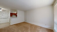 Located at 1402 Fannidella Dr in Carrollton, TX, this single-floor unit offers a practical and efficient layout across its entire floor plan. The combined kitchen and living area forms the heart of the space, with a clear visual connection between the two zones. The galley-style kitchen is well-equipped, featuring warm wood cabinetry lining both walls, a white gas range, a refrigerator, a single-basin sink with ample counter space on either side, and a peninsula that separates the kitchen from the adjoining living area. Decorative wallpaper accents add a classic touch above the upper cabinets. The living area is finished with wall-to-wall carpet in a neutral tan tone, creating a soft and comfortable atmosphere. A window allows natural light to fill the room, complemented by a ceiling-mounted light fixture. The bathroom includes a full tub-and-shower combination with floor-to-ceiling tile surround in a light gray grid pattern, paired with a standard toilet. A dedicated closet provides storage space. Light tile flooring in the kitchen transitions smoothly to carpeting in the living area, defining each zone while maintaining a cohesive overall feel throughout this straightforward and functional single-level unit.