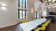 Located at 8560 Second Ave in Silver Spring, Maryland, this thoughtfully designed amenity space offers a welcoming and versatile environment across a single floor. The space is defined by its dramatic double-height ceilings and expansive floor-to-ceiling windows, which flood the interior with natural light and provide views of the surrounding greenery. Warm hardwood-style flooring runs throughout, grounding the open layout with a sense of continuity.

The lounge area is anchored by a comfortable gray sofa, a sleek marble-topped coffee table, and a cozy armchair positioned beside a curved bay window. A bold geometric accent wall in shades of gray, yellow, and white adds a striking visual focal point. Adjacent to the lounge, a game table with yellow accent chairs provides an inviting spot for casual gatherings.

The dining area features a long white communal table surrounded by vibrant yellow chairs, offering ample seating for group events or meetings. A dedicated kitchenette serves the space with modern white cabinetry, a stainless steel refrigerator, a wall-mounted television, and a sink with contemporary fixtures, complemented by stylish pendant lighting. Together, these areas create a cohesive, contemporary space ideal for both social and functional use.
