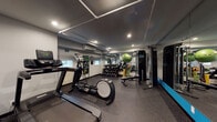 Located at 510 College Ave NE in Grand Rapids, Michigan, this single-floor fitness facility is thoughtfully designed to support a comprehensive range of workout routines. The space is divided into distinct functional zones, creating an organized and efficient environment for users of all fitness levels.

The cardio section is well-stocked with multiple treadmills, a Precor elliptical trainer, and Spinning stationary bikes, all positioned along one side of the room and complemented by a water station for easy hydration access. Recessed lighting and track lighting throughout provide consistent, bright illumination across the entire floor.

The strength training area features a full dumbbell rack with a wide range of weights, a multi-station Precor cable machine, and an adjustable weight bench. Resistance bands, a stability ball, and additional accessories round out the functional fitness offerings. Full-length mirrors line the walls, visually expanding the space while allowing users to monitor their form during exercises.

A wall-mounted split air conditioning unit ensures climate control year-round. The durable rubber flooring throughout is both practical and supportive underfoot. The overall aesthetic is clean and modern, with a neutral gray and white color palette accented by bold teal-blue walls near the entrance, lending the space a polished, professional atmosphere.