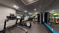 Located at 510 College Ave NE in Grand Rapids, Michigan, this single-floor fitness facility is thoughtfully designed to support a comprehensive range of workout routines. The space is divided into distinct functional zones, creating an organized and efficient environment for users of all fitness levels.

The cardio section is well-stocked with multiple treadmills, a Precor elliptical trainer, and Spinning stationary bikes, all positioned along one side of the room and complemented by a water station for easy hydration access. Recessed lighting and track lighting throughout provide consistent, bright illumination across the entire floor.

The strength training area features a full dumbbell rack with a wide range of weights, a multi-station Precor cable machine, and an adjustable weight bench. Resistance bands, a stability ball, and additional accessories round out the functional fitness offerings. Full-length mirrors line the walls, visually expanding the space while allowing users to monitor their form during exercises.

A wall-mounted split air conditioning unit ensures climate control year-round. The durable rubber flooring throughout is both practical and supportive underfoot. The overall aesthetic is clean and modern, with a neutral gray and white color palette accented by bold teal-blue walls near the entrance, lending the space a polished, professional atmosphere.