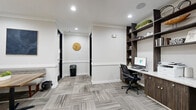 Located in Plano, TX, this business center is a thoughtfully designed, single-floor workspace that offers a professional and welcoming environment. The space is anchored by a dedicated workstation area featuring a built-in dark wood desk with granite-style countertops, two computer stations, a printer, and floor-to-ceiling open shelving adorned with decorative accents and greenery. Ergonomic task chairs complete the functional setup. The lounge area provides comfortable seating with a blue sofa and a booth-style bench surrounding a wooden table, ideal for casual meetings or collaborative work. Two framed abstract artworks lend a refined aesthetic to the walls. Entry is through a striking set of black-framed glass double doors, contributing to the space's modern character. Throughout, patterned gray carpet tiles add texture while maintaining a neutral, professional palette. Recessed lighting illuminates the space evenly, complemented by crown molding details along the ceiling. A wall-mounted clock and clean white walls with contrasting dark trim create a polished, cohesive look. This space is well-suited for productivity, offering both private computer work and relaxed collaborative seating within a compact, efficiently utilized footprint.