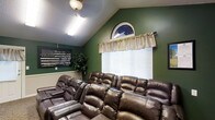 This single-floor space is designed as a dedicated media or lounge room, offering a welcoming and comfortable environment for group gatherings and entertainment. The room's most striking architectural feature is its vaulted ceiling, which adds a sense of openness and height, complemented by a textured ceiling finish. The walls are painted in a deep, rich green, lending the space a warm and immersive atmosphere well-suited for a viewing room.

Natural light enters through a large window adorned with classic white horizontal blinds and a decorative valance in a neutral cream tone. Above the window, a distinctive arched accent window adds an elegant architectural touch. A ceiling-mounted light fixture provides functional overhead illumination throughout the space.

The room is furnished with multiple rows of generously sized brown leather recliner sofas, arranged in a tiered, theater-style layout to ensure clear sightlines for all occupants. This seating arrangement underscores the room's purpose as a communal viewing and relaxation space. Decorative elements include a framed patriotic American flag artwork on the wall and a tall indoor plant, adding character and life to the setting. Wainscoting along the lower portion of the walls provides a refined finishing detail.