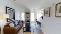 This well-appointed single-floor space offers a thoughtfully designed open layout that balances comfort and functionality. The living area serves as the heart of the space, anchored by a rich brown leather sofa paired with a dark wood coffee table and a textured area rug that adds warmth underfoot. A built-in shelving unit centered between two windows provides both decorative display space and architectural interest, while multiple large windows fitted with classic white blinds flood the room with abundant natural light.

The vaulted ceiling adds a sense of openness and airiness, enhancing the overall spacious feel. Crisp white walls throughout create a clean, neutral backdrop that complements the warm wood tones of the hardwood flooring. Along one wall, a raised bar-height counter with sleek bar stools creates an intimate dining or workspace nook, subtly separated from the main living area. Framed artwork adorns the walls, contributing to an inviting and curated ambiance. Potted greenery and carefully placed decorative accents complete the look, lending a lived-in yet polished character to the space. The overall design strikes a balance between modern simplicity and warm, welcoming comfort.