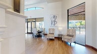Located at 12240 Bella Terra Center Way in Richmond, TX, this single-floor community clubhouse offers a generous and thoughtfully designed living space. Upon entry, a grand glass-paneled door opens into a welcoming foyer featuring warm hardwood-style flooring, crisp white walls, and soaring ceilings that immediately establish a sense of openness and refinement. Contemporary abstract artwork accents the walls, complementing the sophisticated interior aesthetic.

The expansive living area is defined by dramatic vaulted ceilings with exposed wooden beam detailing and large arched clerestory windows that flood the space with natural light. Multiple seating arrangements — including tufted wingback chairs, a plush sectional sofa, and accent tables — create distinct zones for relaxation and conversation. A mounted flat-screen television and a classic fireplace serve as focal points along one wall, flanked by an elegant ornate sideboard.

A billiards table anchors the entertainment zone, positioned beneath a striking geometric chandelier. Floor-to-ceiling windows line the perimeter, offering panoramic views of the outdoor pool area and surrounding landscaping. Mixed flooring of hardwood and travertine tile adds visual texture throughout. The overall ambiance is upscale yet inviting, blending modern design sensibilities with warm, transitional elements.