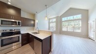 This single-story unit is defined by its striking vaulted ceilings, which lend a sense of openness and volume to the main living area. The open-concept layout connects the kitchen and living space seamlessly, with light wood-tone flooring running throughout and an abundance of natural light streaming in through large, multi-pane windows that frame views of surrounding greenery.

The kitchen is well-appointed with dark espresso cabinetry, white quartz-style countertops, and a full suite of stainless steel appliances, including a refrigerator, electric range, built-in microwave, and dishwasher. A peninsula with pendant lighting overhead serves as a natural divider between the kitchen and living area. A dedicated in-unit laundry space houses a full-size washer and dryer with wire shelving above for additional storage.

The bedroom features soft carpeting and a window with natural light, alongside access to a spacious walk-in closet outfitted with wire shelving and hanging rods on multiple walls. The bathroom is clean and functional, featuring a full soaking tub with shower, a dark-cabinetry vanity with white countertop, light wood-look flooring, and neutral wall tones throughout.