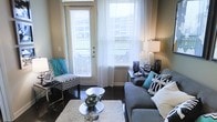 This single-floor space showcases a thoughtfully designed living room that maximizes its compact footprint through strategic furniture placement and a cohesive color scheme. The room is anchored by large windows dressed in sheer white curtains and horizontal blinds, allowing generous natural light to fill the space while maintaining privacy. A glass-paneled exterior door further enhances the sense of openness and provides access to the outdoors.

The living area features dark hardwood flooring that contrasts elegantly with a plush white area rug, creating a defined and inviting seating zone. A gray sectional sofa dressed with teal and graphic black-and-white accent pillows serves as the focal point, complemented by a chevron-patterned accent chair adorned with a teal throw. A sleek, round glass coffee table adds a modern touch without overwhelming the space.

Decorative elements include table lamps, framed artwork, and carefully curated accessories in coordinating tones of teal, black, and silver. The overall ambiance is contemporary and welcoming, balancing style with functionality in a well-lit, efficiently arranged environment.