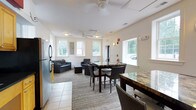 This single-floor community room is designed to accommodate both relaxed gatherings and seated group activities. The space is divided into two functional zones: a lounge area and a dining area, unified by neutral-toned walls and textured carpeting throughout.

The lounge area is furnished with dark wicker-style sofas and armchairs arranged around a glass-topped coffee table, creating a comfortable setting for informal socializing. Ceiling fans overhead provide air circulation, while wall-mounted sconce lighting adds a warm ambiance. Multiple double-hung windows frame views of the surrounding neighborhood, filling the space with generous natural light.

The dining area features two substantial tables with stone-look surfaces, accompanied by wicker dining chairs with upholstered seats. Additional windows along the dining side further enhance the brightness of the room.

A kitchenette is accessible from the main space, equipped with warm wood cabinetry, dark granite countertops, and a stainless steel sink—providing convenient amenities for gatherings and events. Tile flooring in the kitchenette transitions seamlessly to the carpeted main area. An exterior door with glass panes offers direct access to the outdoors, completing the room's functional and welcoming design.